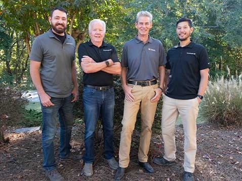 4 men in polo shirts standing in a row by a small river